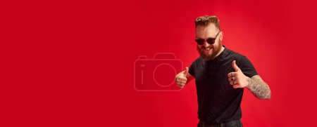 Foto de Retrato de hombre con estilo barbudo en gafas de sol alegremente posando sobre fondo rojo estudio. Concepto de estilo de vida, estilo escocés, moda, música, diversión y alegría. Banner. Copiar espacio para anuncio - Imagen libre de derechos