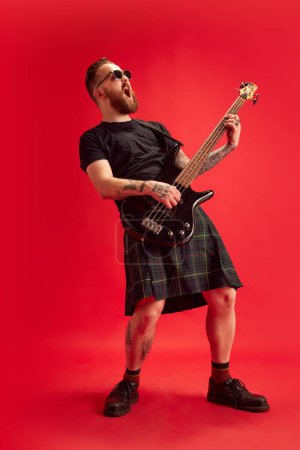 Foto de Hombre barbudo en gafas de sol y falda, falda escocesa tocando la guitarra sobre fondo rojo estudio. Rock and roll. Concepto de estilo de vida, estilo escocés, moda, música, diversión y alegría, anuncio - Imagen libre de derechos