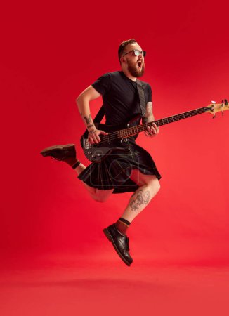 Foto de Hombre barbudo en gafas de sol y falda, falda escocesa tocando la guitarra sobre fondo rojo estudio. Músico emocional. Concepto de estilo de vida, estilo escocés, moda, música, diversión y alegría, anuncio - Imagen libre de derechos