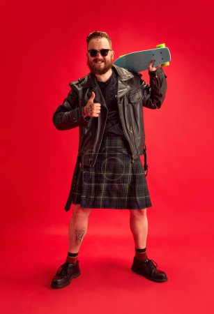 Photo for Brutal, stylish, bearded man in leather jacket, sjirtk, kilt and singlsees rjumping with skateboard against red studio background. Concept of lifestyle, scottish style, fashion, music, fun and joy, ad - Royalty Free Image