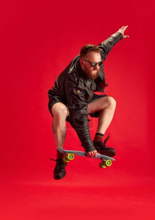 Photo for Brutal, stylish, bearded man in leather jacket, sjirtk, kilt and singlsees rjumping with skateboard against red studio background. Concept of lifestyle, scottish style, fashion, music, fun and joy, ad - Royalty Free Image