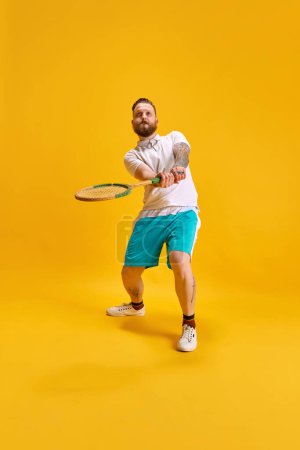 Téléchargez les photos : Image pleine longueur de l'homme barbu en vêtements de sport posant avec raquette de tennis sur fond de studio jaune. Concentration, jeu. Concept de sport, force, mode, émotions, mode de vie, publicité - en image libre de droit