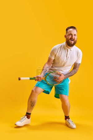Photo for Full-length image of bearded man in sportswear playing tennis racket like guitar against yellow studio background. Imagination and fun. Concept of sport, strength, fashion, emotions, lifestyle, ad - Royalty Free Image