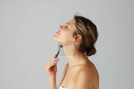 Photo for Beautiful young woman taking doing massage of neck with roller against grey studio background. Rejuvenating treatment. Concept of natural beauty, cosmetology and dermatology, skincare, cosmetics, ad - Royalty Free Image