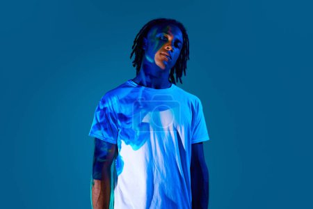 Photo for Creative portrait of african young man with dread with neon light reflection on body standing against blue background in neon light. Concept of art, fashion, modern style, cyberpunk, futurism, ad - Royalty Free Image