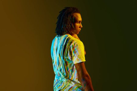 Téléchargez les photos : Portrait latéral de jeune homme africain avec réflexion numérique colorée sur le corps debout sur fond vert dégradé dans la lumière au néon. Concept d'art, mode, style moderne, cyberpunk, futurisme, publicité - en image libre de droit