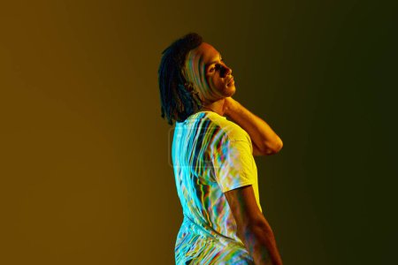 Foto de Retrato de vista lateral del joven africano con reflexión digital colorida sobre el cuerpo de pie sobre el fondo verde degradado en luz de neón. Concepto de arte, moda, estilo moderno, cyberpunk, futurismo, anuncio - Imagen libre de derechos