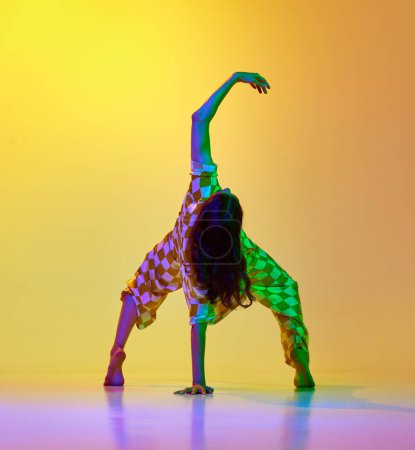 Foto de Imagen dinámica de una joven flexible y artística que baila sobre un fondo amarillo anaranjado degradado con luz de neón. Concepto de estilo de baile moderno, hobby, arte, performance, estilo de vida, anuncio - Imagen libre de derechos
