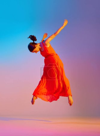 Foto de Mujer joven talentosa y hermosa en vestido rojo elegante bailando contra el fondo multicolor degradado en luz de neón. Concepto de estilo de baile moderno, hobby, arte, performance, estilo de vida, anuncio - Imagen libre de derechos
