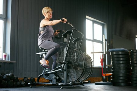 Foto de Mujer hermosa madura en entrenamiento de ropa deportiva en el gimnasio, haciendo ejercicios en bicicleta estacionaria. Manteniendo el cuerpo en tono. Concepto de estilo de vida activo, deportivo y saludable, fitness, cuidado corporal, anuncio - Imagen libre de derechos