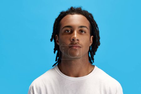 Téléchargez les photos : Portrait de jeune homme africain avec des dreads en t-shirt blanc regardant la caméra sur fond bleu studio. Regard calme et détendu. Concept de jeunesse, émotions humaines, mode de vie, mode, publicité - en image libre de droit