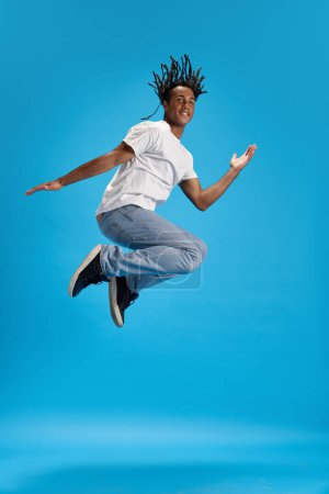 Foto de Imagen de cuerpo entero del joven africano con rastas en ropa casual saltando sobre fondo azul del estudio. Me siento positivo. Concepto de juventud, emociones humanas, estilo de vida, moda, anuncio - Imagen libre de derechos