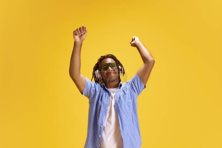 Téléchargez les photos : Jeune homme en lunettes de soleil et vêtements décontractés écoutant de la musique dans des écouteurs et dansant sur fond de studio jaune. Concept de jeunesse, émotions humaines, mode de vie, mode, expressions faciales, publicité - en image libre de droit
