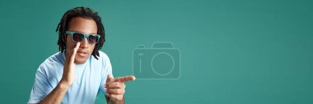 Photo for Whispering secrets. Young guy in sunglasses holding hand to mouth and pointing with finger against green studio background. Concept of youth, human emotions, lifestyle, fashion, facial expressions, ad - Royalty Free Image