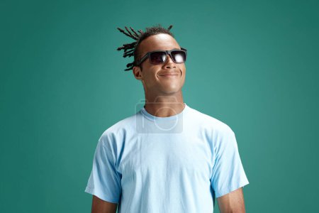 Foto de Retrato de un joven guapo, alegre y positivo con gafas de sol de pie y sonriendo sobre el fondo verde del estudio. Concepto de juventud, emociones humanas, estilo de vida, moda, expresiones faciales, anuncio - Imagen libre de derechos