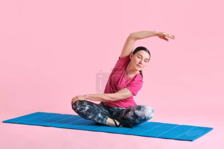 Photo for Beautiful mature woman sitting on fitness matt in lotus pose and stretching against pink studio background. Concept of sport, healthy lifestyle, fitness, body care, wellness, ad - Royalty Free Image