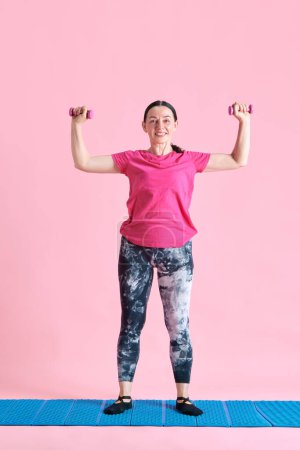 Foto de Sonriente, maduro, entrenamiento de mujer deportiva, haciendo ejercicios con pesas sobre fondo de estudio rosa. Concepto de deporte, estilo de vida saludable, fitness, cuidado del cuerpo, bienestar, anuncio - Imagen libre de derechos