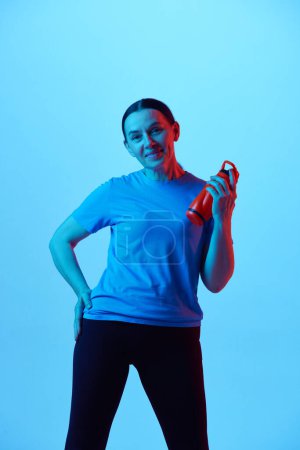 Téléchargez les photos : Portrait de belle femme mature avec corps en forme debout avec bouteille d'eau sur fond de studio bleu dans la lumière au néon. Concept de sport, mode de vie sain, fitness, soins du corps, bien-être, publicité - en image libre de droit