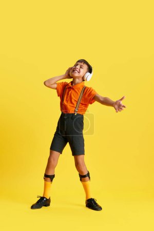 Photo for Full-length of little boy, child in stylish clothes listening to music in headphones against yellow studio background. Concept of childhood, kids emotions, fashion, hobby, ad - Royalty Free Image