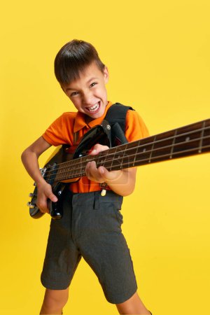 Photo for Emotional boy, child in stylish clothes playing guitar against yellow studio background. Music lifestyle and education. Concept of childhood, kids emotions, fashion, hobby, ad - Royalty Free Image