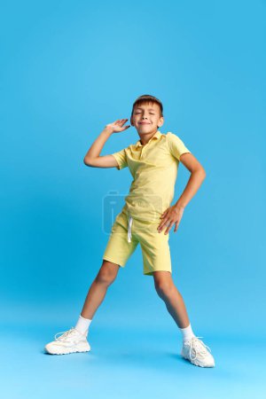 Téléchargez les photos : Pleine longueur de gai petit garçon, enfant en vêtements décontractés posant, dansant sur fond de studio bleu. Détendez-vous. Concept d'enfance, émotions des enfants, vacances, été, plaisir et joie, annonce - en image libre de droit