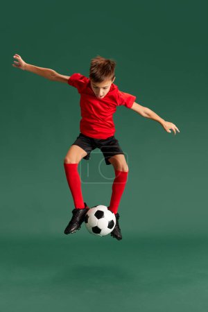 Foto de Futuro futbolista. Niño pequeño, niño en ropa deportiva posando, jugando con la pelota contra el fondo del estudio verde. Concepto de infancia, emociones de los niños, estilo de vida deportivo, acción, hobby, anuncio - Imagen libre de derechos