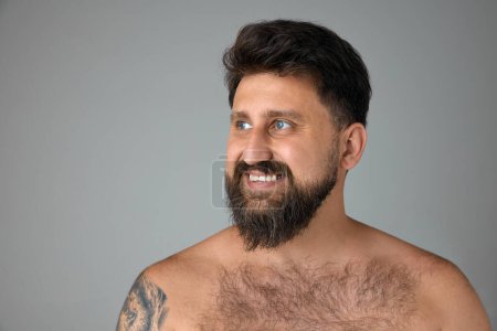 Photo for Handsome, smiling, mature, bearded man with blue eyes standing shirtless, looking away against grey studio background. Spa. Concept of mens beauty, skin care, cosmetology, health and wellness. - Royalty Free Image