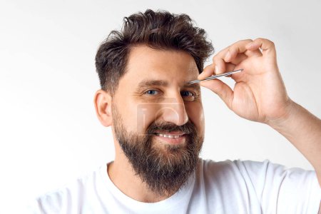 Photo for Bearded mature man taking care after appearance, using tweezers for good looking brows against white studio background. Concept of mens beauty, skin care, cosmetology, health and wellness. - Royalty Free Image