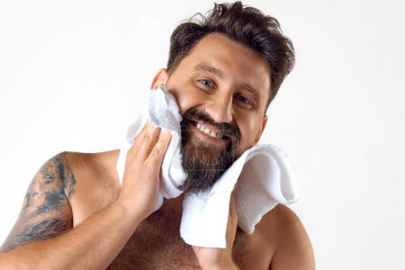Photo for Portrait of handsome, smiling, bearded shirtless man with towel on shoulder washing face against white studio background. Concept of mens beauty, skin care, spa, cosmetology, health and wellness. - Royalty Free Image