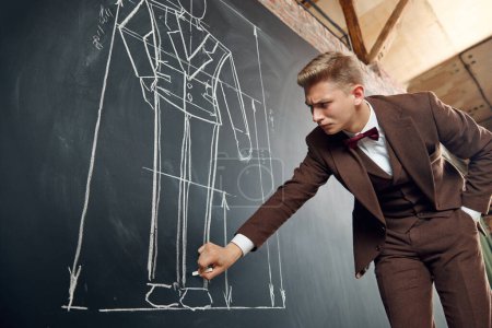 Photo for Concentrated young man in brown classic suit, fashion designer making drawings on blackboard, creating new clothes. Concept of fashion, profession, creativity, occupation, hobby, business - Royalty Free Image