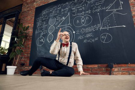 Photo for Young man, teacher, scientist sitting by blackboard with scientific calculations and formulas, finding out solution. Concept of education, science, profession and occupation, knowledge - Royalty Free Image