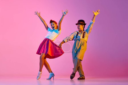 Foto de Niños emocionales y elegantes, niño y niña en ropa retro bailando lindy hop contra fondo de estudio rosa en luz de neón. Concepto de infancia, hobby, estilo de vida activo, performance, arte, moda - Imagen libre de derechos