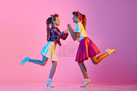 Foto de Chicas hermosas y elegantes, niños en ropa retro bailando lindy hop contra fondo de estudio rosa en luz de neón. Concepto de infancia, hobby, estilo de vida activo, performance, arte, moda - Imagen libre de derechos
