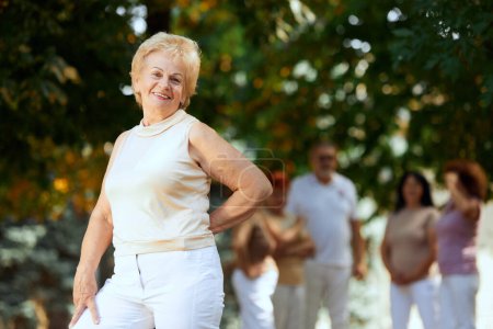Téléchargez les photos : Concentrez-vous sur les femmes âgées positives en vêtements blancs debout dans le parc par une chaude journée ensoleillée. Des gens flous sur le fond. Concept de sport et de santé, mode de vie actif, âge, bien-être, corps, soins - en image libre de droit