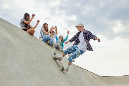 Téléchargez les photos : Les amis, les teengaers se réunissent au skate park pour des loisirs actifs. Garçon en vêtements casucl skateboard sur la rampe. Concept de culture de la jeunesse, sport, dynamique, extrême, passe-temps, action et mouvements, amitié - en image libre de droit
