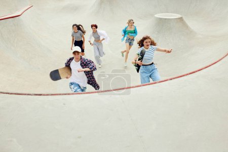 Téléchargez les photos : Groupe d'adolescents en vêtements décontractés, garçons et filles qui courent avec du skate sur une rampe de skateboard. Activité et plaisir. Concept de culture de la jeunesse, sport, dynamique, extrême, passe-temps, action et mouvements, amitié - en image libre de droit