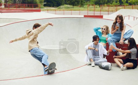 Foto de Amigos alegres, adolescentes reunidos en el parque de skate, divertirse, montar en el patín, hablar. Chico haciendo acrobacias. Concepto de cultura juvenil, deporte, dinámica, extrema, afición, acción y movimientos - Imagen libre de derechos