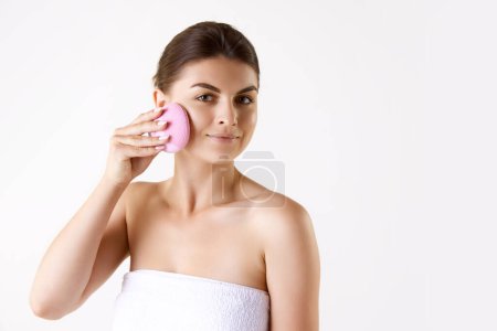 Photo for Attractive woman after shower, in towel cleansing face with special face tool against white studio background. Concept of female beauty, skin care, cosmetology and cosmetics, health, ad - Royalty Free Image