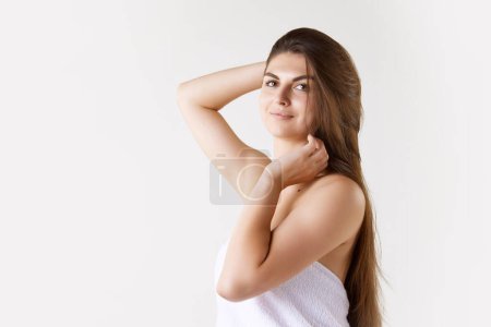 Téléchargez les photos : Portrait de belle jeune femme brune aux longs cheveux raides, debout dans la serviette sur fond de studio blanc. Concept de beauté féminine, soins de la peau, cosmétologie et cosmétique, santé, publicité - en image libre de droit