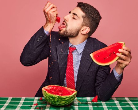 Photo for Man in a suit eating watermelon against pink background. Green checkered tablecloth. Juicy drops. Concept of food, creativity, party, summer, health. Pop art photography. Copy space for ad - Royalty Free Image