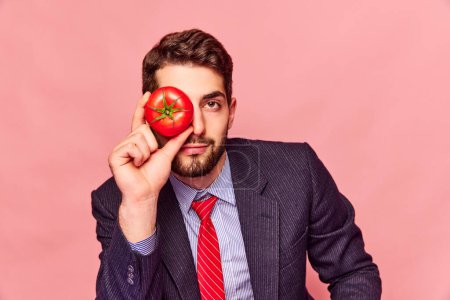 Téléchargez les photos : Homme barbu en costume classique et cravate rouge tenant la tomate près des yeux et regardant la caméra sur fond rose rose. Concept de nourriture, créativité, santé. Photographie pop art. Espace de copie pour la publicité - en image libre de droit