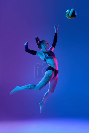 Foto de Longitud completa de la joven mujer deportiva, jugador de voleibol en movimiento, golpeando la pelota en un salto contra fondo de estudio azul en luz de neón. Concepto de deporte profesional, competición, salud, hobby, anuncio - Imagen libre de derechos