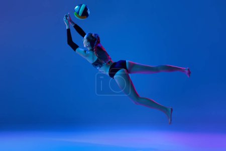 Foto de Imagen dinámica de la mujer joven durante el juego, jugando al voleibol, golpeando la pelota contra el fondo del estudio azul en luz de neón. Concepto de deporte profesional, competición, salud, hobby, acción, anuncio - Imagen libre de derechos