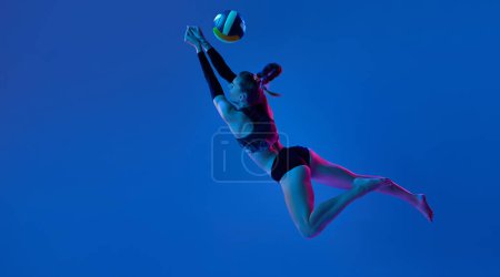 Téléchargez les photos : Athlète professionnelle féminine, jeune femme jouant au volley-ball, en mouvement frappant la balle contre fond bleu studio sous la lumière du néon. Concept de sport professionnel, compétition, santé, passe-temps, action, publicité - en image libre de droit
