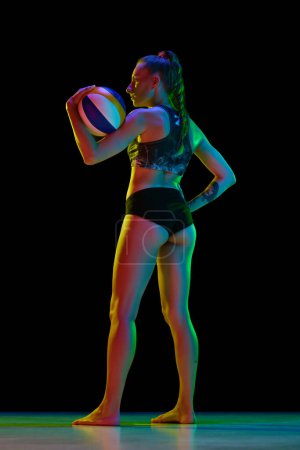 Téléchargez les photos : Vue latérale complète de la jeune femme, joueuse de volley-ball en tenue de sport debout avec balle sur fond de studio noir au néon. Concept de sport professionnel, compétition, santé, action, publicité - en image libre de droit