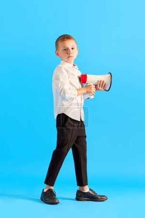 Foto de Pequeño colegial, niño con camisa blanca y pantalones negros sosteniendo megáfono sobre fondo azul del estudio. Noticias. Concepto de infancia, educación, escuela, emociones infantiles, redes sociales, anuncios - Imagen libre de derechos