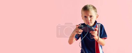 Foto de Retrato de un niño serio, niño con ropa deportiva, jugando a la consola sobre fondo rosa del estudio. Videojuegos en línea. Concepto de infancia, emoción infantil, estilo de vida, diversión y alegría, anuncio. Banner - Imagen libre de derechos