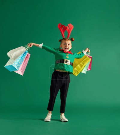 Téléchargez les photos : Petit garçon en costume vert d'elfe tenant de nombreux sacs à provisions sur fond de studio vert. Les courses de Noël. Concept de vacances d'hiver, émotions des enfants, mode, shopping, enfance, publicité - en image libre de droit