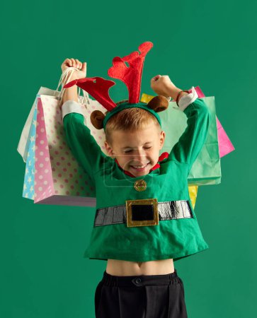 Téléchargez les photos : Petit garçon souriant en costume vert d'elfe tenant de nombreux sacs à provisions sur fond de studio vert. Des cadeaux. Concept de vacances d'hiver, émotions des enfants, mode, shopping, enfance, publicité - en image libre de droit