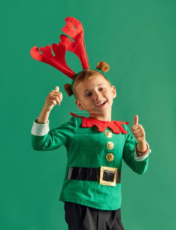 Téléchargez les photos : Portrait pour petit garçon, enfant heureux en costume d'elfe posant joyeusement sur fond de studio vert. Concept de vacances d'hiver, émotions des enfants, mode, shopping, enfance, publicité - en image libre de droit